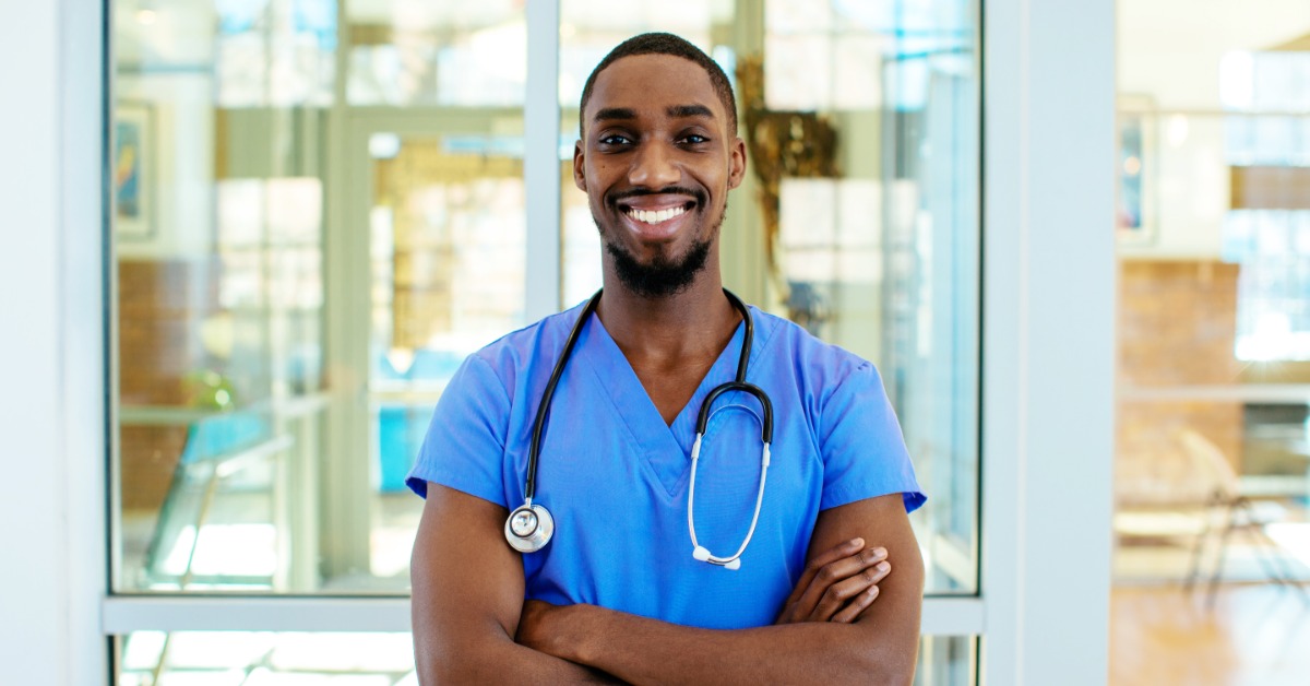 Smiling healthcare professional in blue scrubs with stethoscope stands with arms crossed in bright hospital corridor.