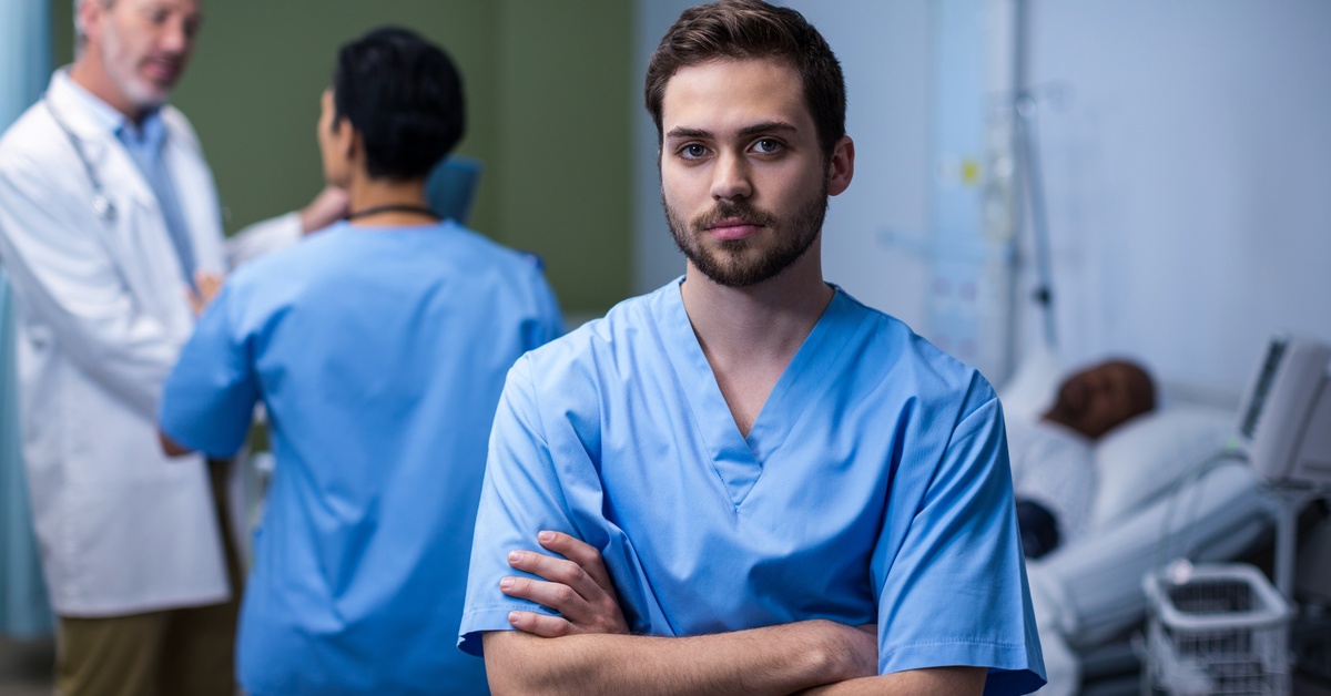 A man wearing blue scrubs stands with his arms crossed over his chest. A patient lies in a bed behind him.