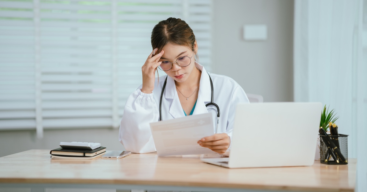 A woman wearing a white lab coat and glasses with a stethoscope around her neck looking at a document as she sits at a desk.