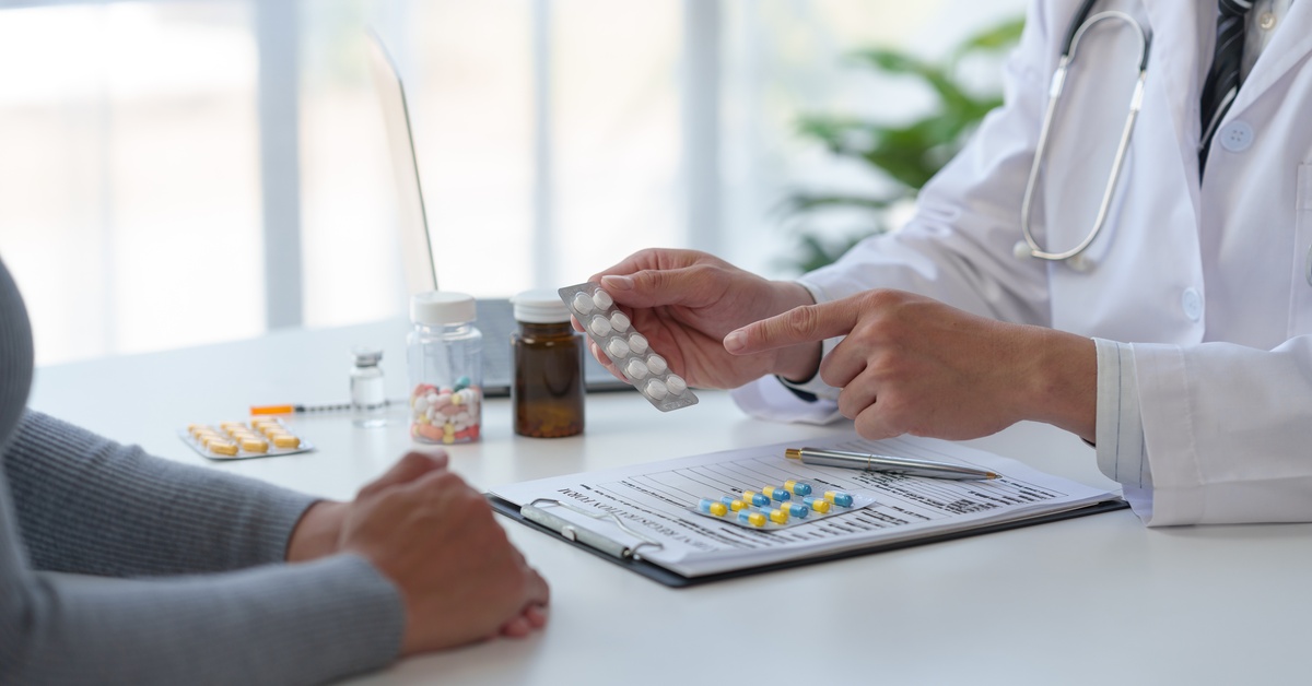 A person wearing a white doctor's coat and a stethoscope points to a blister pack of pills in front of another person.