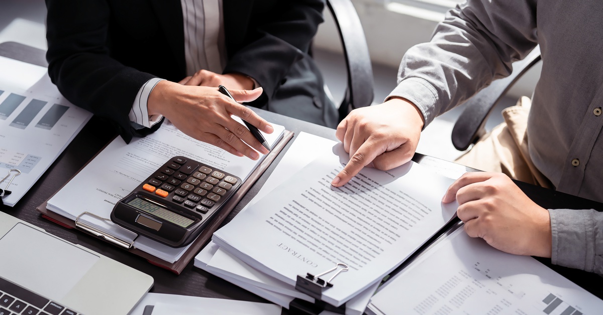 Two people sit at a desk holding documents and a calculator. One person is pointing at something on the document.