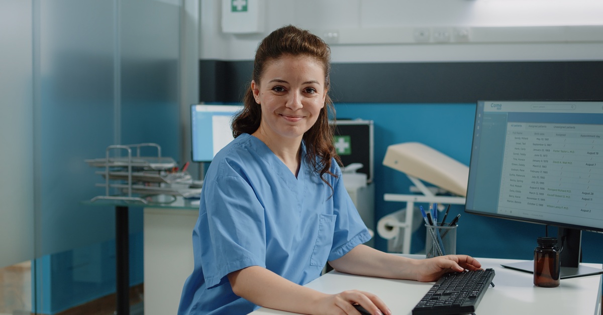 A woman wearing blue scrubs sits at a computer with her hand on the mouse and smiles in a medical setting.
