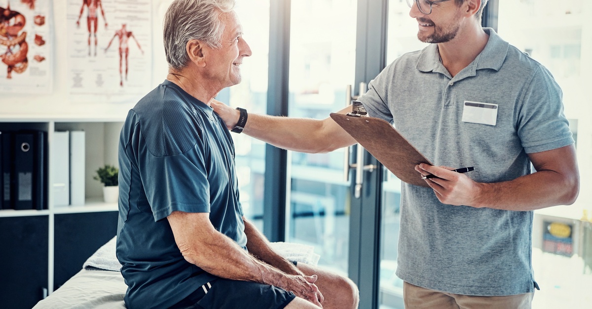 A man holding a clipboard puts a hand reassuringly on a patient's shoulder. The patient is sitting on an exam table.