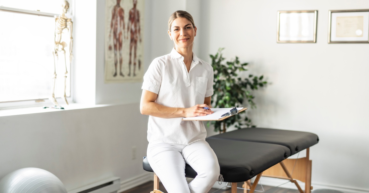 A woman holds a clipboard as she sits on an exam table in a room with a model of a skeleton and posters of the muscular structure.