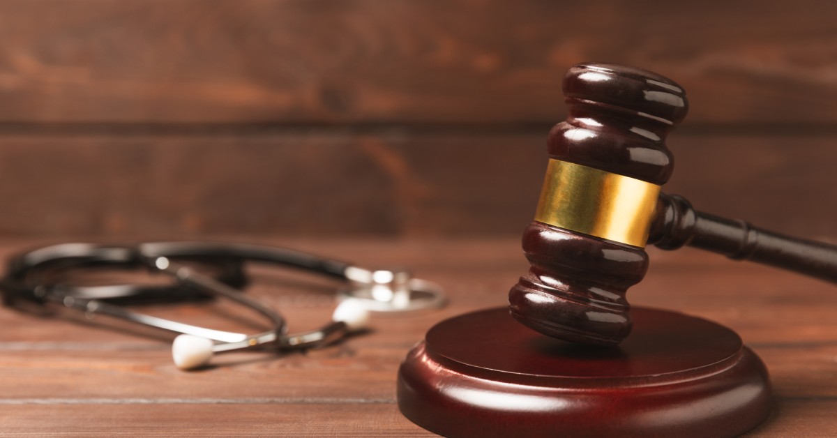 A close-up of a wooden gavel resting on a polished base. A blurred stethoscope sits in the background on a wooden surface.